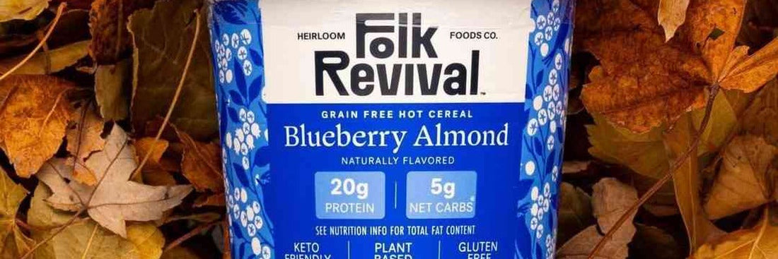A Folk Revival Blueberry Almond hot cereal cup lying in autumn leaves, the label showing 20 g protein and 5 g net carbs.