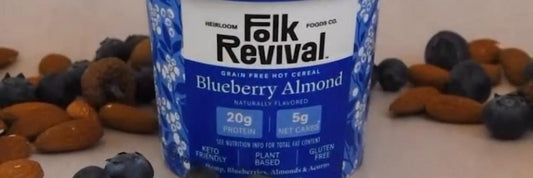 A Folk Revival Blueberry Almond hot cereal cup surrounded by fresh blueberries and almonds.