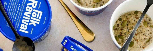 Small bowls of Folk Revival hot cereal topped with seeds, beside a cup and its lid, seen from above.